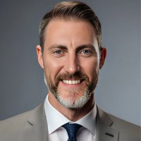 A man with short brown hair and a beard smiles at the camera. He is wearing a light gray suit, a white shirt, and a dark tie. The background is a plain, muted gray.