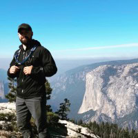 A person in hiking gear stands on a rocky outcrop with a scenic view of a vast canyon and a large rock formation under a clear blue sky. Sparse trees are visible around the rocky area.
