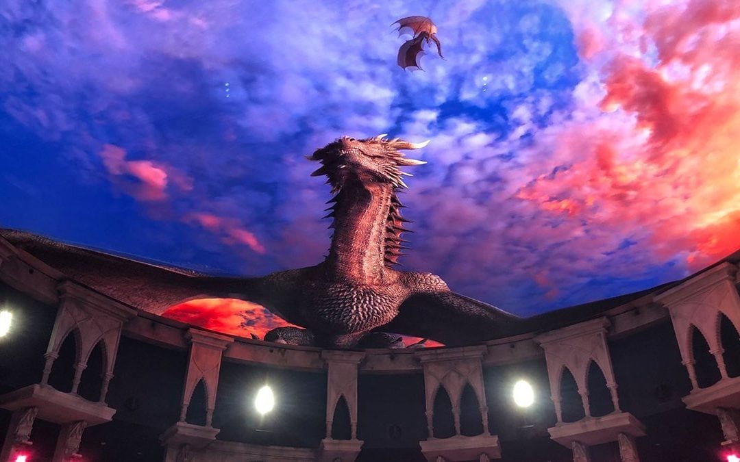 A large, detailed dragon emerges on an immersive, circular screen amidst a vibrant, fiery sky. A smaller dragon flies gracefully above. The setting echoes an arena with arches and lit torches—it's like stepping into a fantastical show at the Ron Clark Academy.