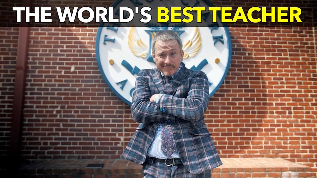 A person in a plaid suit, reminiscent of Ron Clark's charismatic style, stands confidently with arms crossed in front of a brick building featuring a large crest. The text above reads, "The World’s Best Teacher.